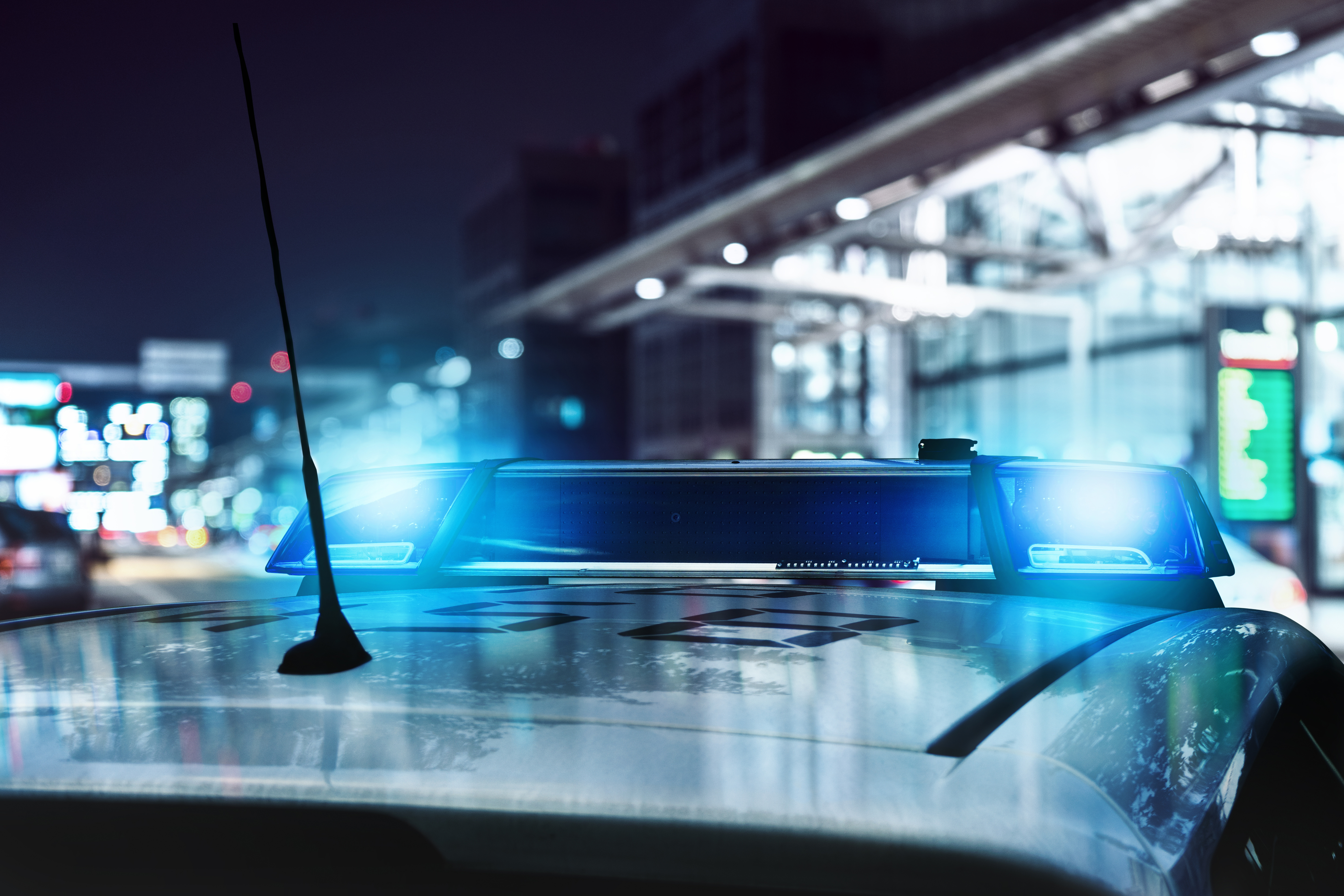 The roof of a patrol car can be seen in the foreground. The blue light is switched on. A bus station can be seen in the background on the right, a larger road on the left. It is dark outside.