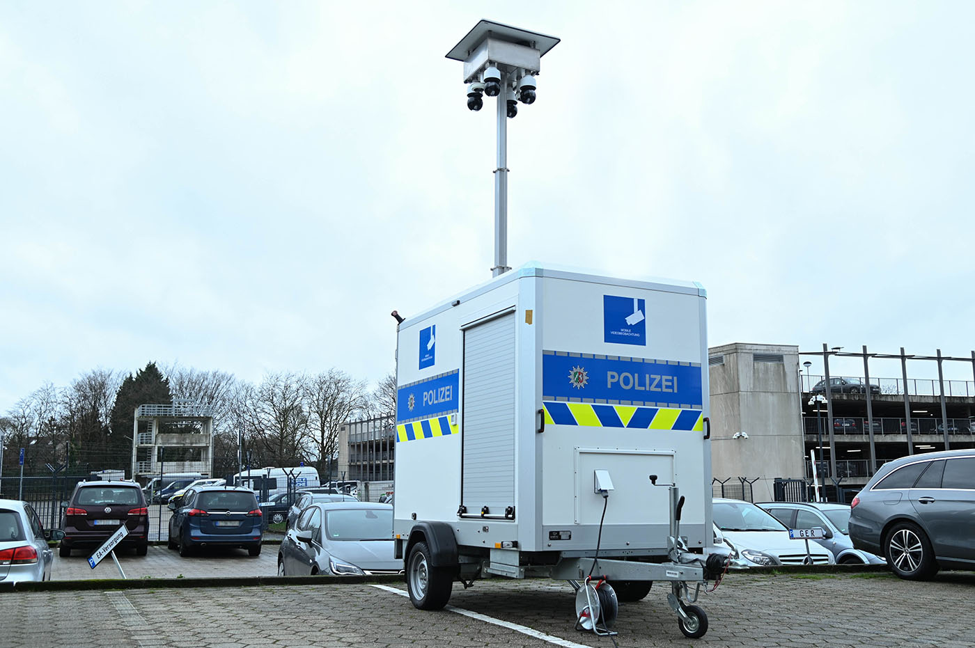 Ein Fahrzeuganhänger der Polizei NRW, auf dem ein ausfahrbarer Kameramast befestigt ist, steht auf einem Parkplatz.