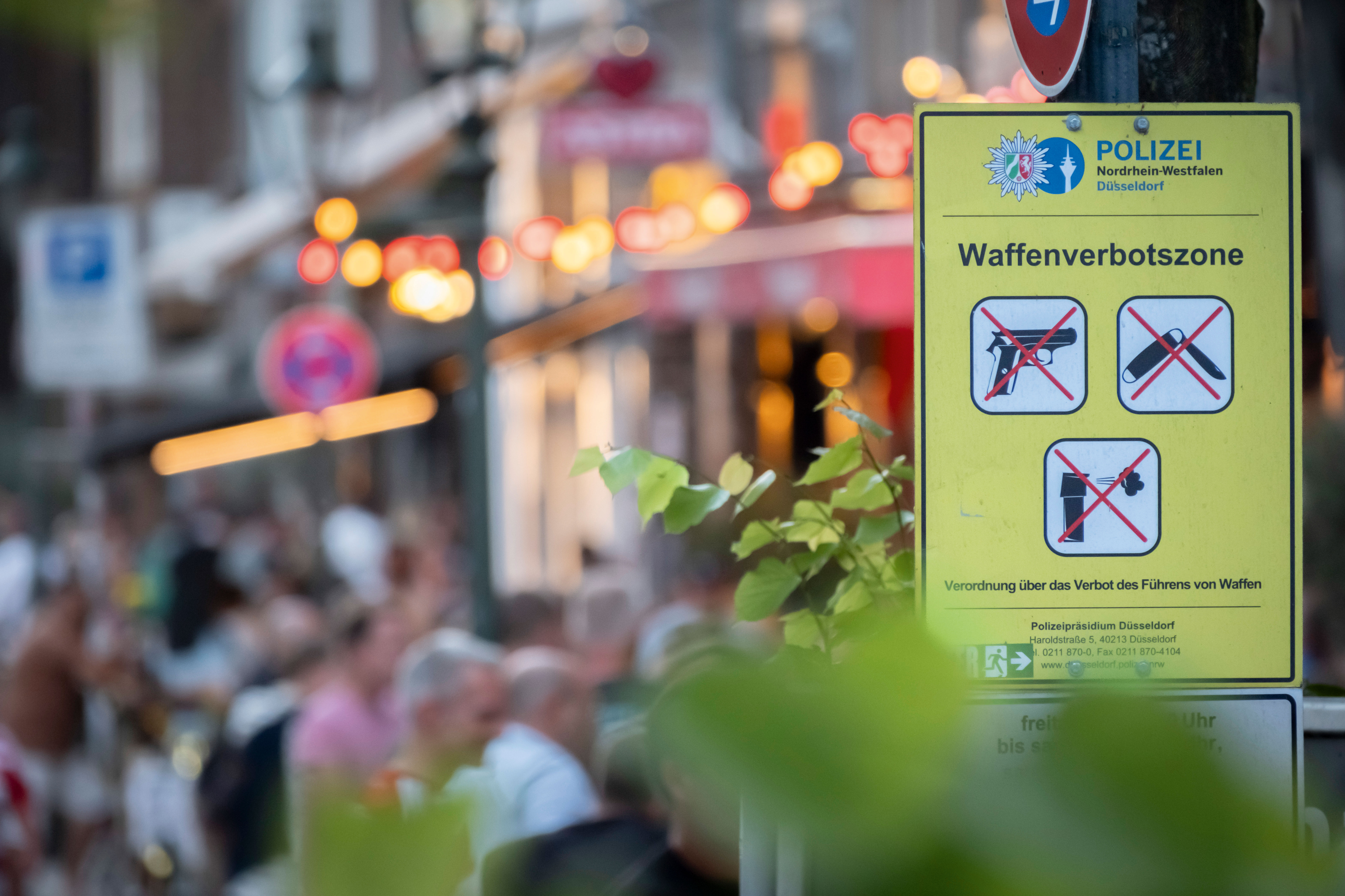 Im Hintergrund ist verschwommen ein Abschnitt der Düsseldorfer Altstadt zu sehen. Davor steht ein gelbes Schild mit der Aufschrift "Waffenverbotszone". Auf dem Schild sind Bilder verschiedener Waffen zu sehen, die mit einem Roten X durchgestrichen sind.