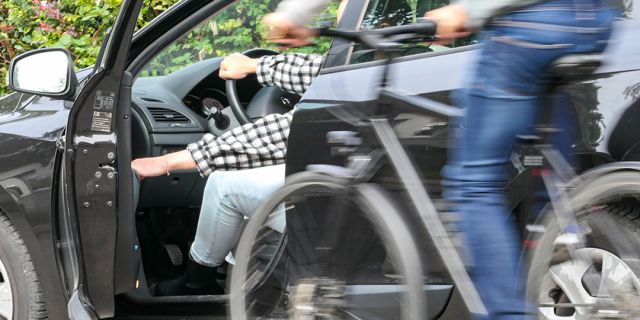 Ein Mann fährt auf einem Fahrrad an einem geparkten Wagen vorbei, dessen Fahrerin gerade ihre Tür öffnet. Die Polizei rät: Vor dem Öffnen einer Autotür immer auch den rückwärtigen Verkehr beachten. Dabei hilft der „Holländische Griff“.