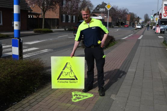 Auf dem Bild steht Polizist Markus Tappe in Greven auf einem Radweg, in der Hand hält er eine Schablone zum Aufsprühen eines Zeichens auf den Asphalt. Das Zeichen zeigt einen Radfahrer in Form eines Geistes und unter dem Emblem steht "Falsche Seite!". Damit sollen Radfahrer darauf hingewiesen werden, die richtige Straßenseite zu benutzen. 