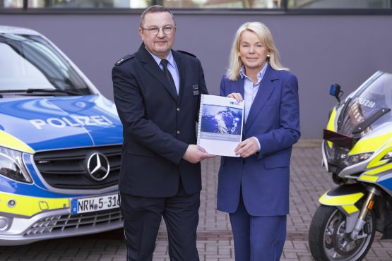 Rainer Kollburg (stellvertretender Leiter der Direktion Verkehr) und Polizeipräsidentin Friederike Zurhausen präsentieren den aktuellen Verkehrsunfallbericht 2024. Links ist ein Streifenwagen zu sehen, rechts ein Polizeimotorrad.
