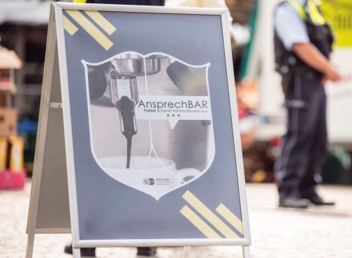 Aufsteller mit Logo der "AnsprechBAR-Veranstaltungsreihe" - auf dem Logo ist eine Kaffeemaschine zu erkennen, aus der Kaffee in eine Tasse läuft. Dazu der Schriftzug "AnsprechBAR - die Polizei schenkt Informationen aus"